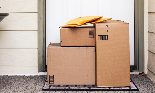 Stacked on a doormat, three cardboard boxes and two padded envelopes sit at a doorstep. The scene conveys anticipation and delivery.