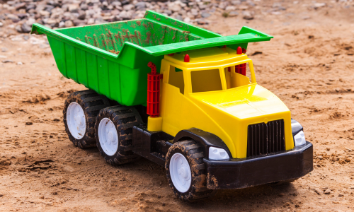 A colorful plastic toy dump truck with a yellow cab and green bed sits on sandy ground, evoking a playful, outdoor, and imaginative atmosphere.