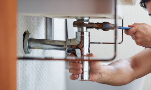 A plumber uses a wrench to fix a silver pipe under a sink, focusing intently. The scene conveys concentration and hands-on repair work.