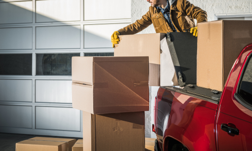 A person wearing a brown jacket and yellow gloves loads cardboard boxes from a red truck in front of a white garage, suggesting a moving or delivery scene.